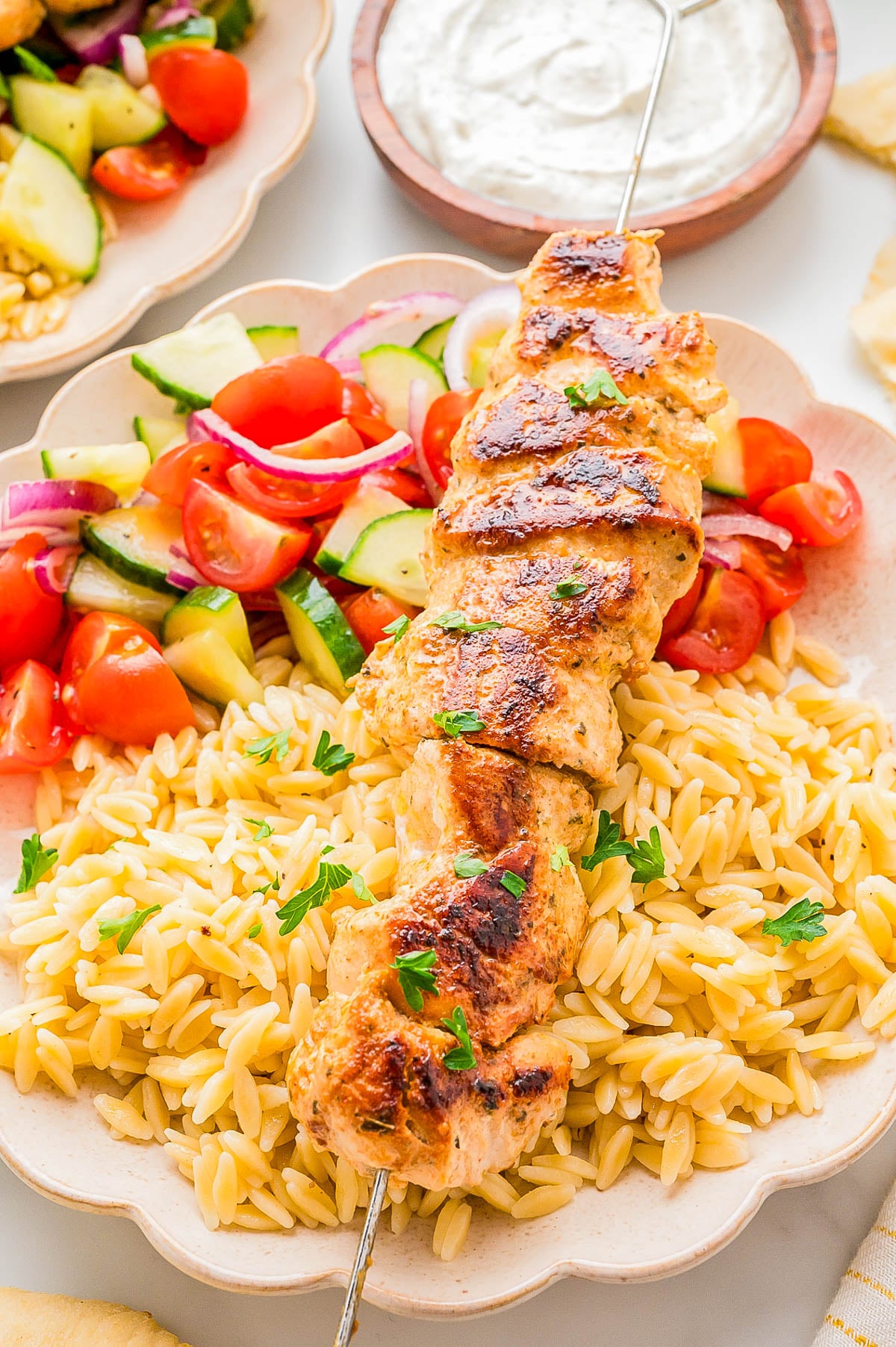 Grilled chicken skewer served over orzo pasta with a side of cucumber and tomato salad; a bowl of white dipping sauce is in the background.
