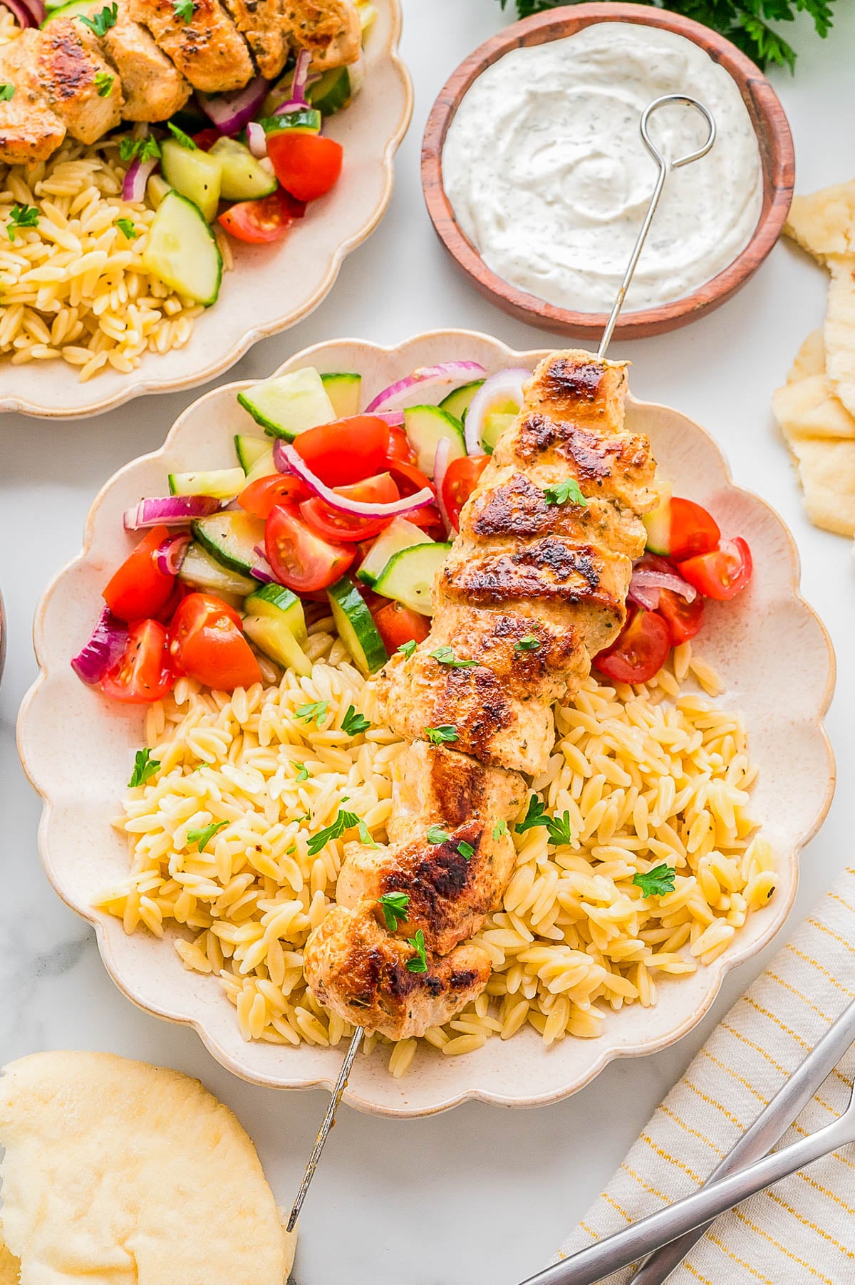 A plate with grilled chicken skewer, orzo pasta, cucumber-tomato salad with red onions, a pita slice, and a bowl of white sauce on the side.