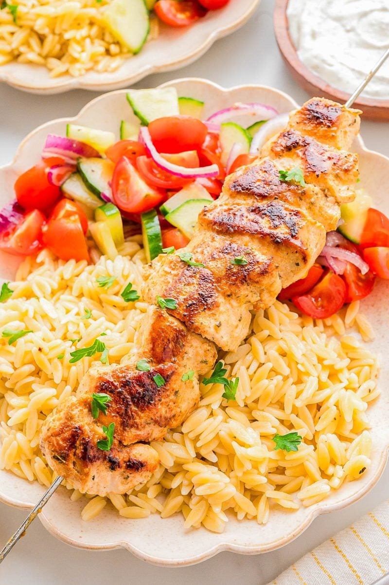 Grilled chicken skewer served over orzo pasta with a side of cucumber, tomato, and red onion salad on a beige plate.