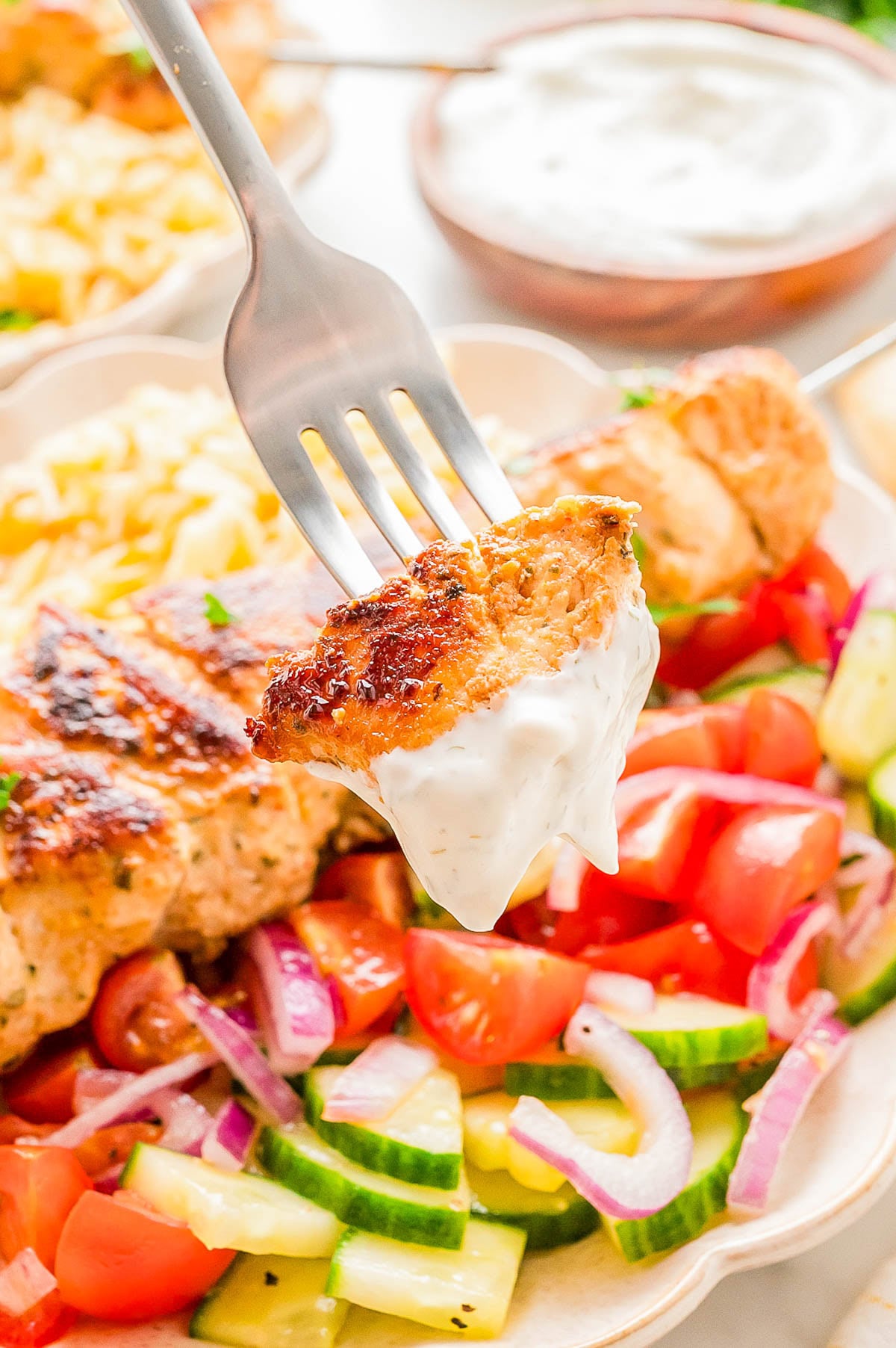 A fork holds a piece of grilled chicken dipped in white sauce above a plate with chopped salad, grilled chicken, and vegetables.