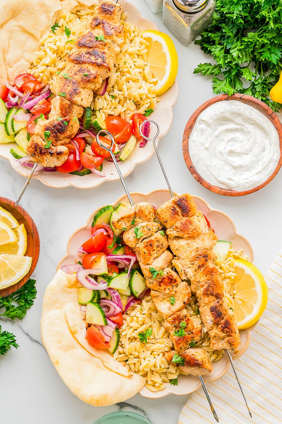 Two plates with grilled chicken skewers, orzo pasta, cucumber tomato salad, pita bread, lemon slices, and a bowl of tzatziki sauce on a white surface.