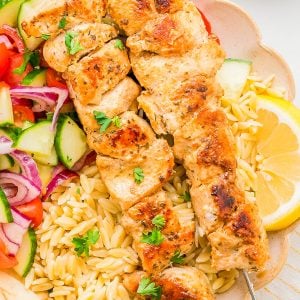 Two grilled chicken skewers served over orzo pasta with a side of cucumber-tomato salad, lemon wedge, and pita bread on a plate.