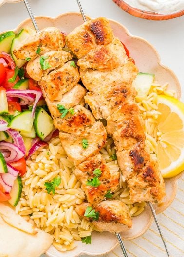 Two grilled chicken skewers served over orzo pasta with a side of cucumber-tomato salad, lemon wedge, and pita bread on a plate.