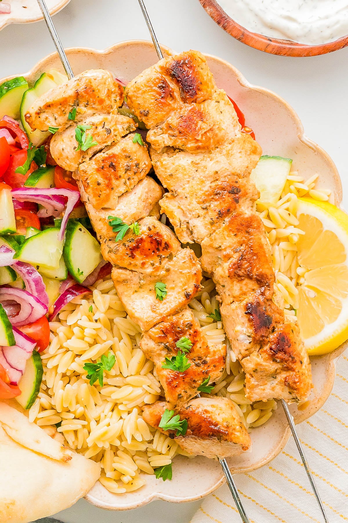 Two grilled chicken skewers served over orzo pasta with a side of cucumber-tomato salad, lemon wedge, and pita bread on a plate.