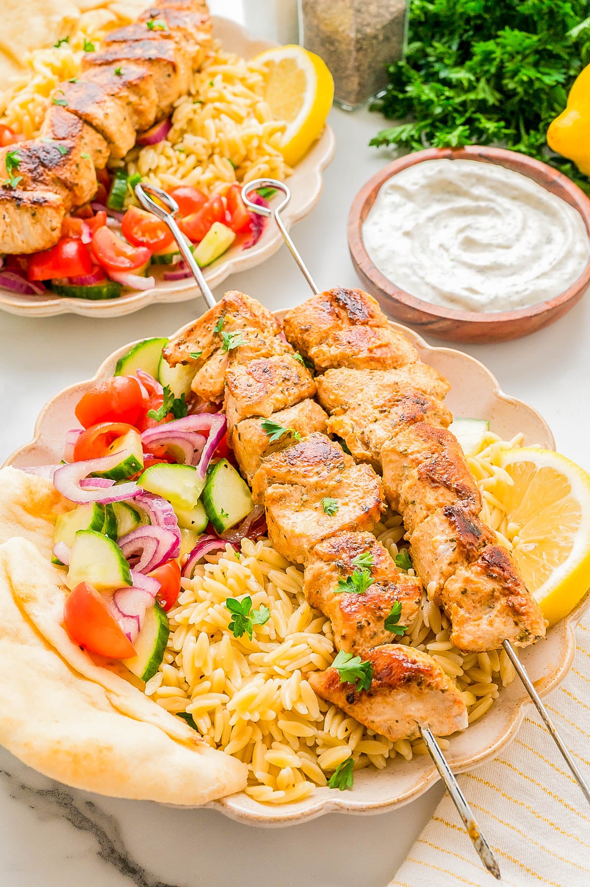 Two skewers of grilled chicken served with orzo, sliced cucumbers, tomatoes, red onions, lemon, pita bread, and a side of creamy white sauce on a plate.