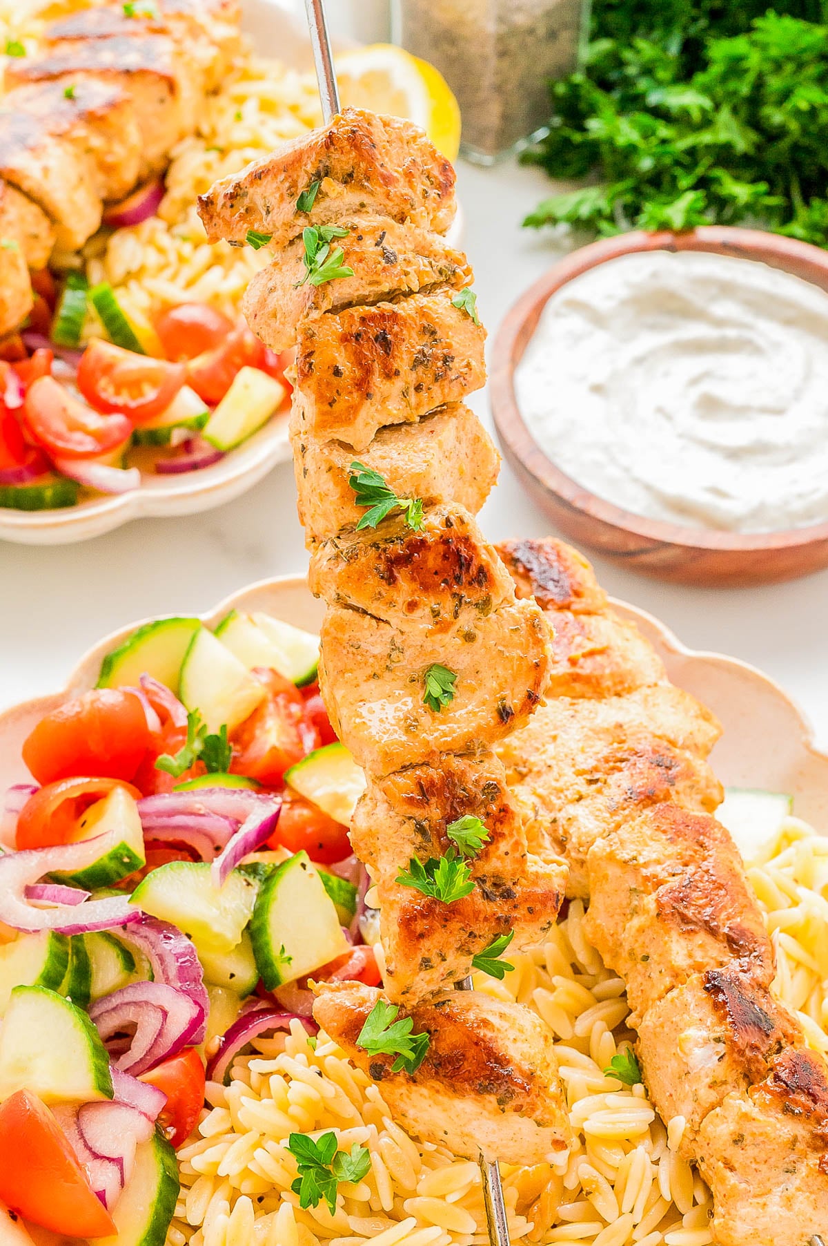 A plate of grilled chicken skewers served with orzo pasta, a side salad with tomatoes, cucumbers, and onions, and a bowl of white dipping sauce in the background.