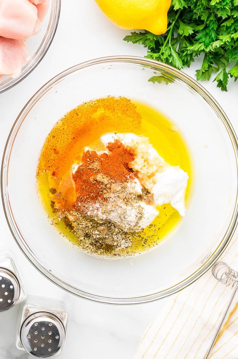 A glass bowl contains yogurt, olive oil, minced garlic, mixed spices, and herbs on a white surface, with parsley, a lemon, and chicken pieces nearby.