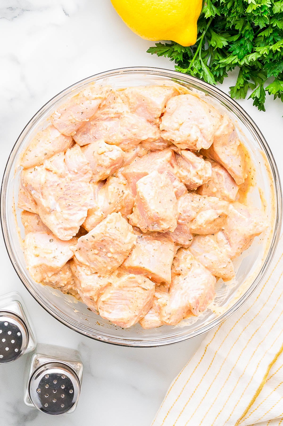 A glass bowl filled with raw, marinated chicken pieces sits on a marble surface next to a lemon, parsley, salt and pepper shakers, and a striped cloth.