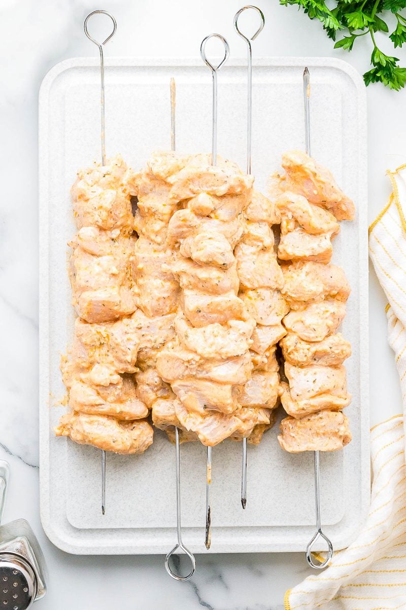 Raw marinated chicken pieces threaded onto metal skewers, arranged on a white cutting board with parsley and a striped towel nearby.