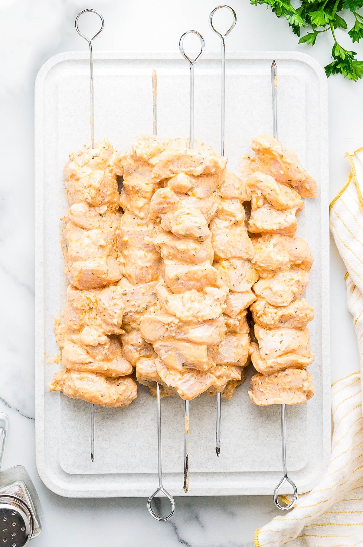Raw marinated chicken pieces threaded onto metal skewers, arranged on a white cutting board with parsley and a striped towel nearby.