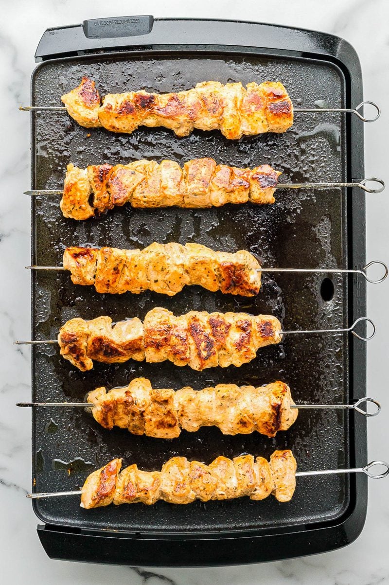 Five grilled chicken skewers on metal sticks cooking on an electric griddle, with visible grill marks and sizzling oil.
