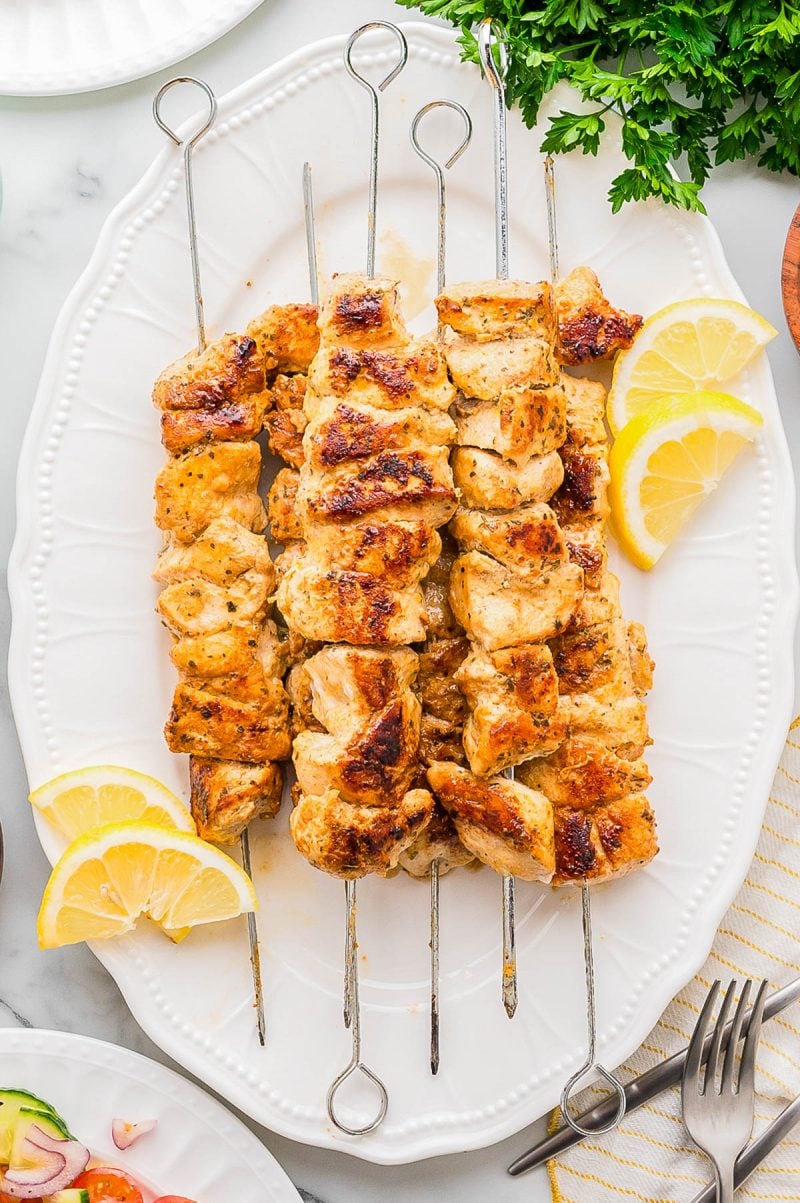 Four grilled chicken skewers are arranged on a white platter, garnished with lemon wedges and fresh parsley.