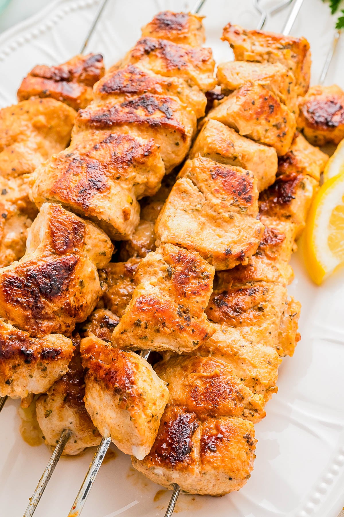 Grilled chicken skewers with a golden-brown crust served on metal sticks, arranged on a white plate with a lemon wedge.