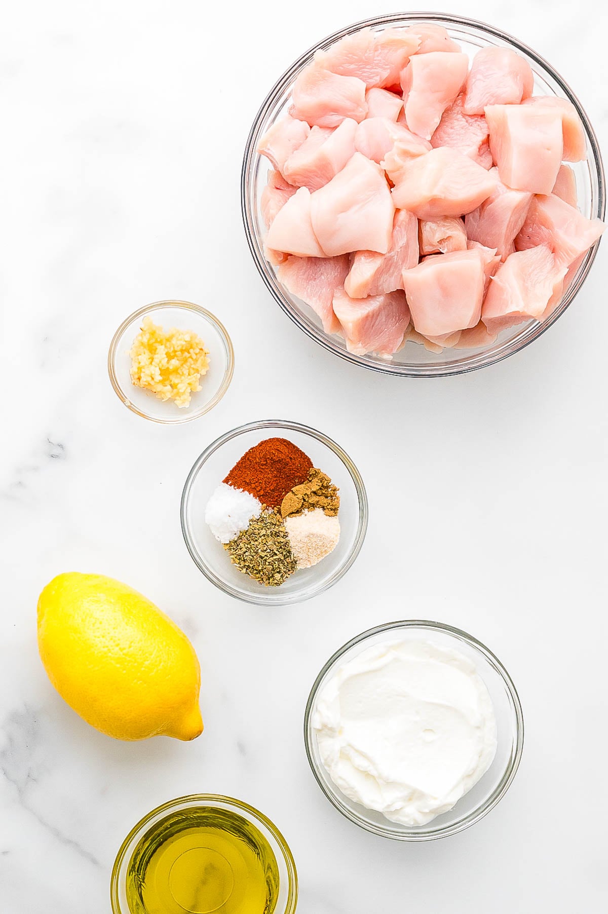 Bowls of raw chicken pieces, minced garlic, mixed spices, yogurt, and olive oil arranged on a white surface next to a whole lemon.