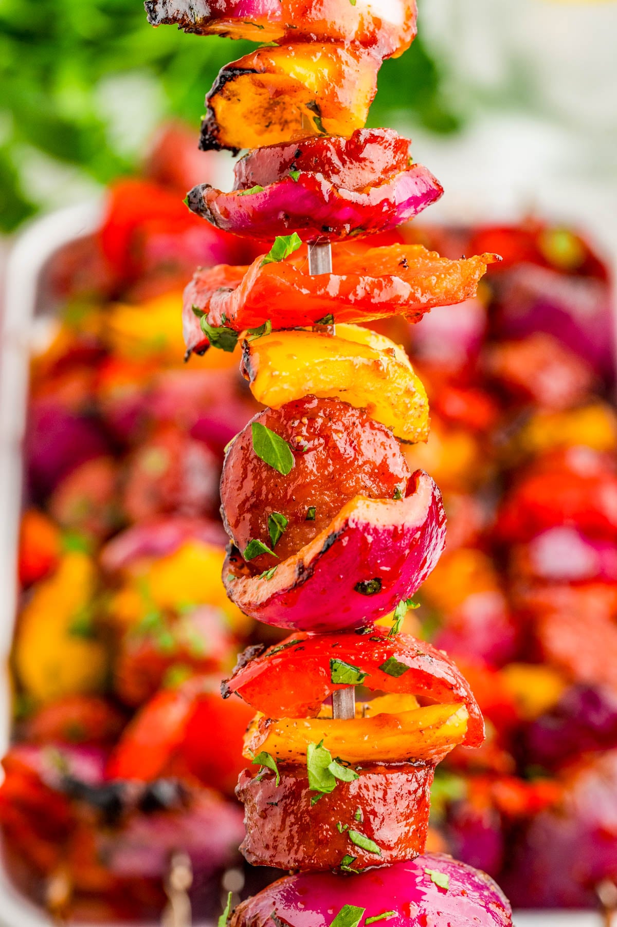 A close-up of a skewer with grilled sausage, red onion, and bell peppers, garnished with chopped herbs.