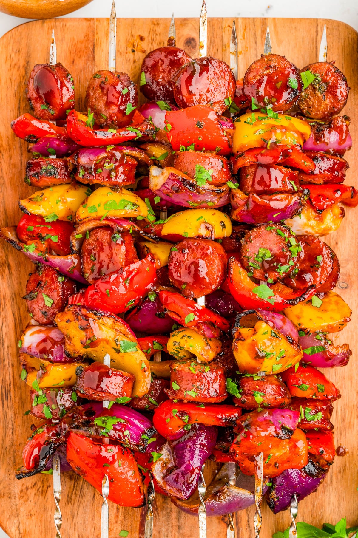 Grilled skewers with sausage slices, red and yellow bell peppers, and red onions, garnished with chopped herbs, arranged on a wooden cutting board.