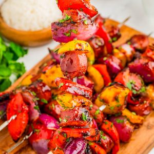Grilled vegetable skewers with red onions, bell peppers, and herbs are arranged on a wooden board, with a bowl of rice and fresh cilantro in the background.