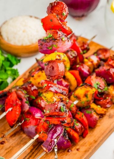 Grilled vegetable skewers with red onions, bell peppers, and herbs are arranged on a wooden board, with a bowl of rice and fresh cilantro in the background.