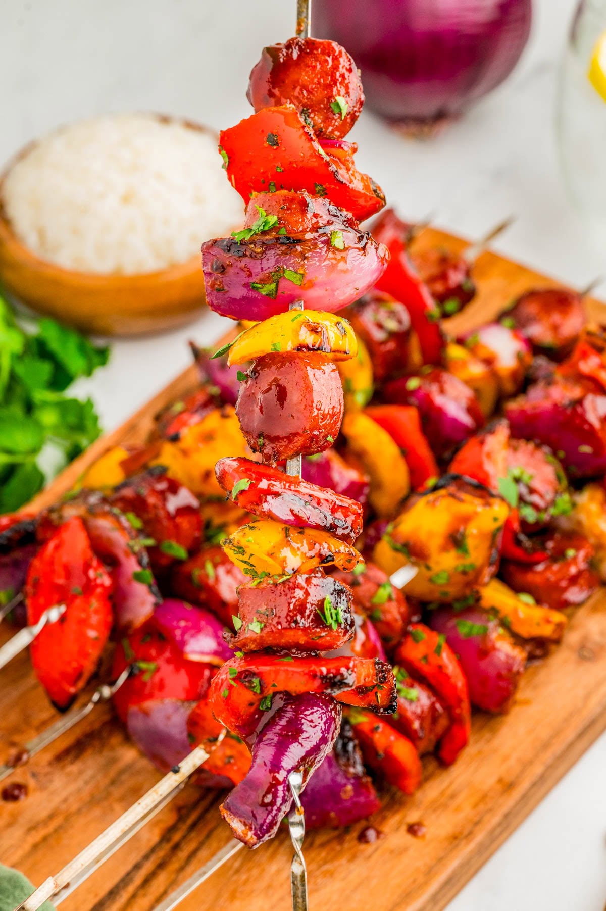 Grilled vegetable skewers with red onions, bell peppers, and herbs are arranged on a wooden board, with a bowl of rice and fresh cilantro in the background.