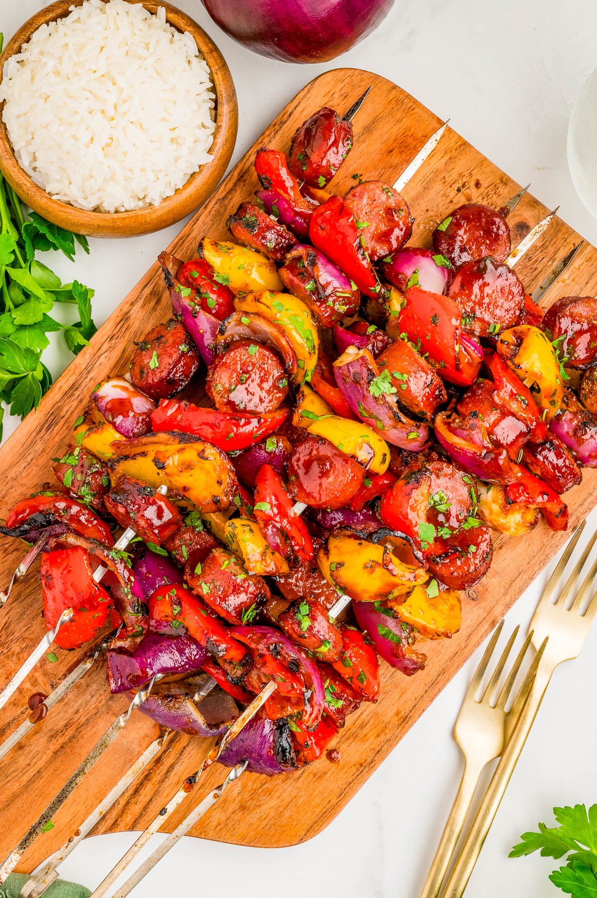 Grilled skewers with sausage, red and yellow bell peppers, and red onions on a wooden board, garnished with herbs, beside a bowl of white rice and fresh parsley.