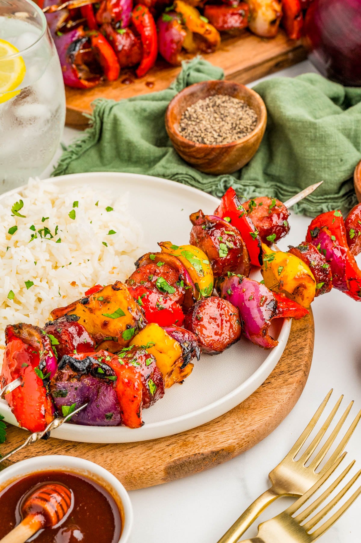 A plate with grilled vegetable and sausage skewers, white rice, a small bowl of sauce, gold utensils, and a glass of water with lemon.