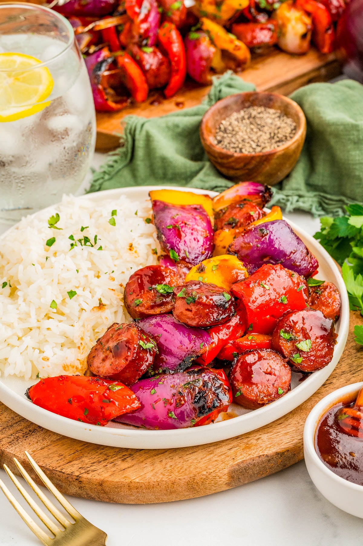 A plate of white rice served with grilled sausage, red onions, and bell peppers, garnished with herbs. A glass of water with lemon and a small bowl of pepper are nearby.