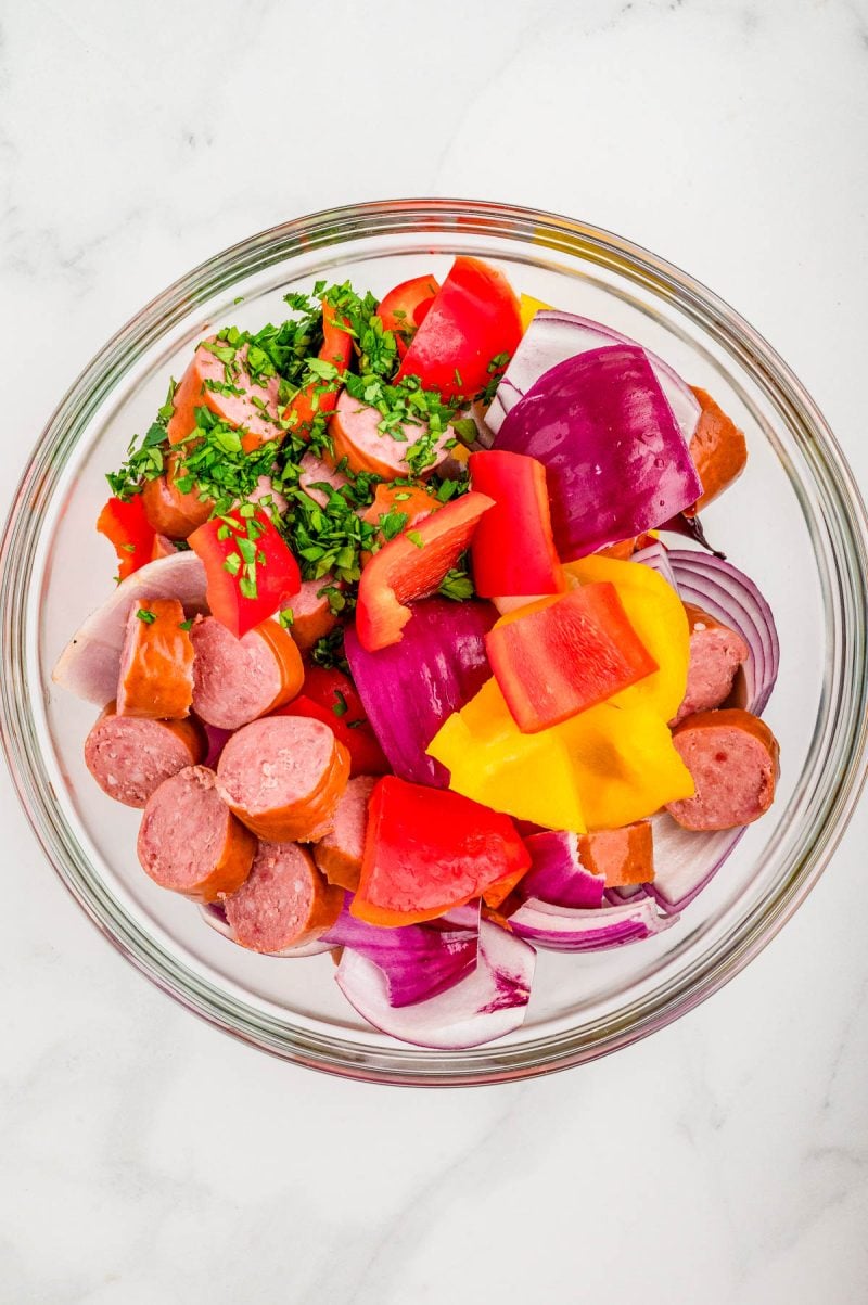 A glass bowl containing sliced sausage, chopped red and yellow bell peppers, red onion, and fresh herbs on a white surface.
