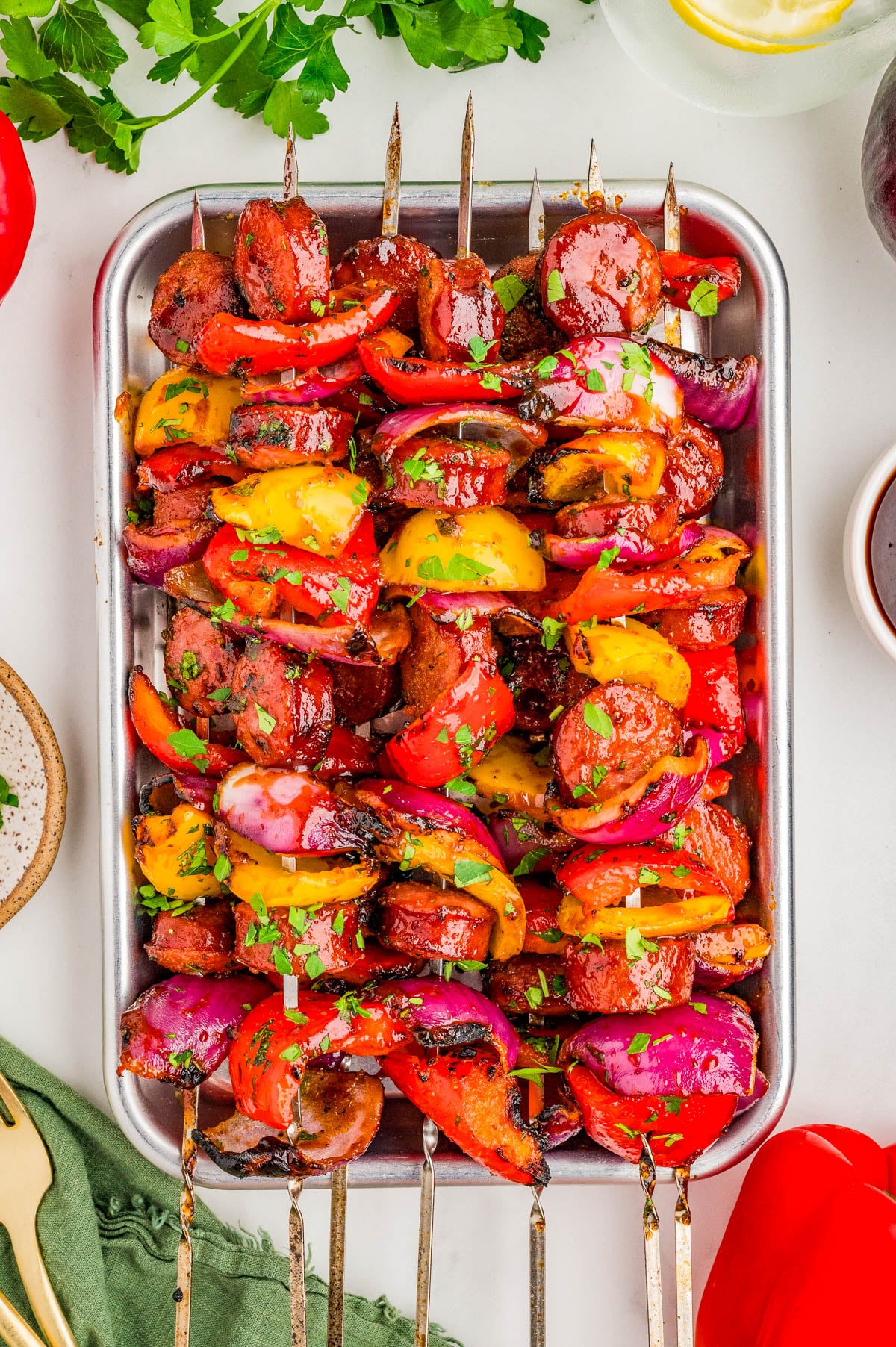 Grilled skewers with sausage, red onions, and red and yellow bell peppers garnished with chopped herbs on a metal tray.