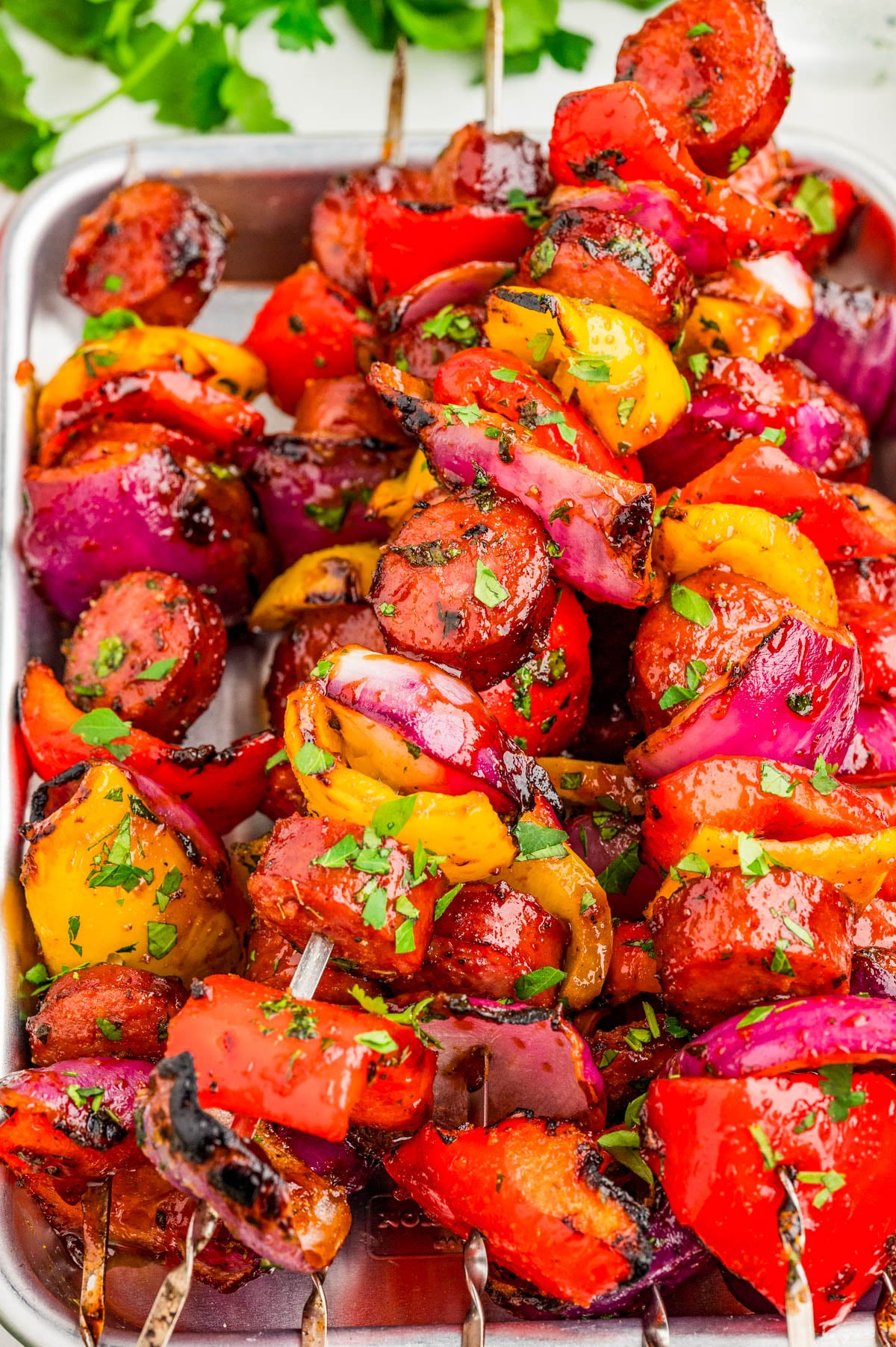 Grilled skewers with sausage, red onions, red and yellow bell peppers, garnished with chopped herbs, served on a metal tray.