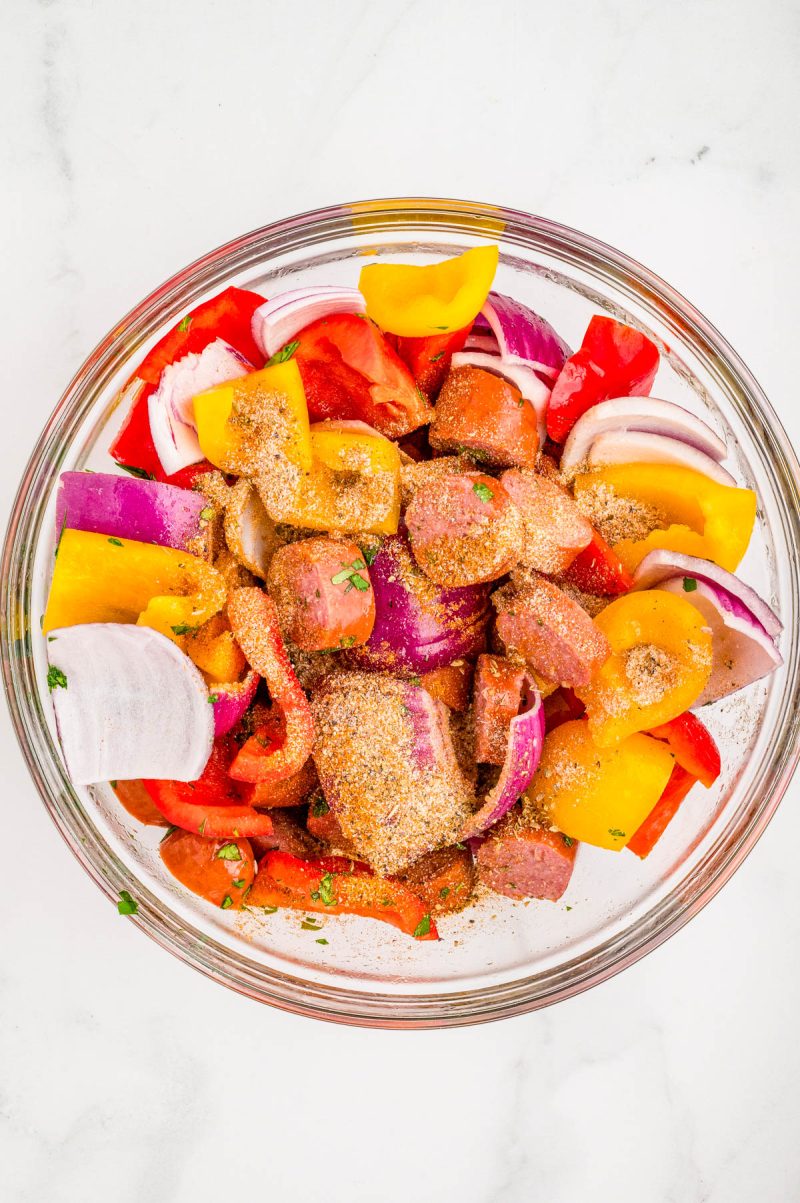 A glass bowl filled with chopped sausage, red and yellow bell peppers, red onion, and spices on a white surface.