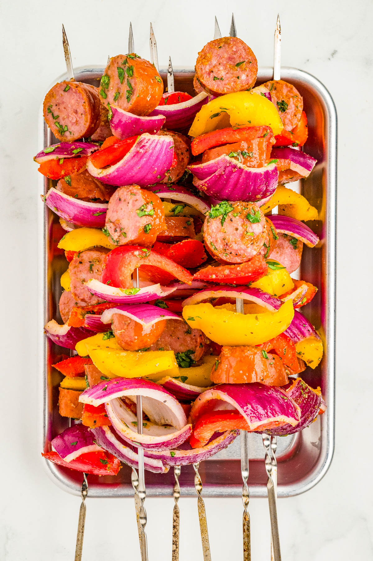Metal skewers with sliced sausage, red onion, yellow bell pepper, and red bell pepper arranged in a metal tray on a white surface.