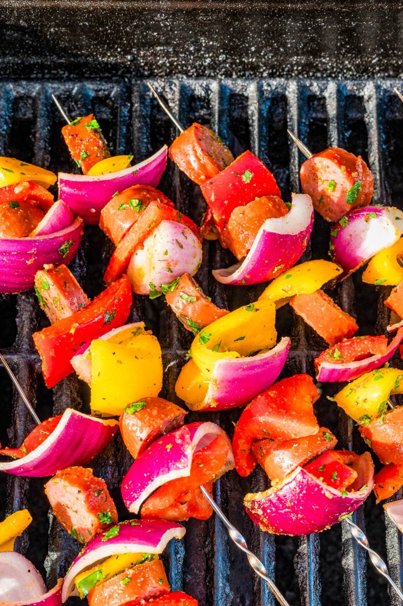 Colorful skewers with sausage, red onion, and yellow bell pepper grilling on a barbecue.