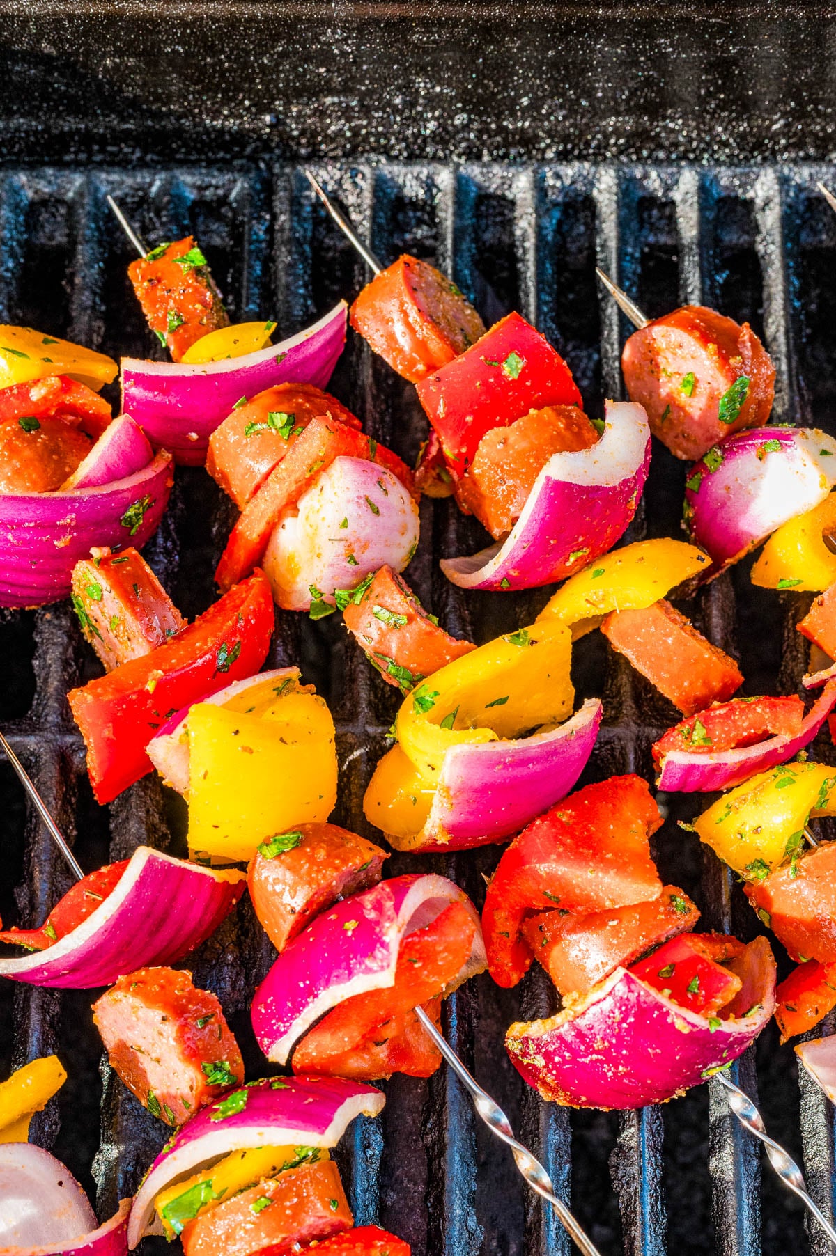 Colorful skewers with sausage, red onion, and yellow bell pepper grilling on a barbecue.