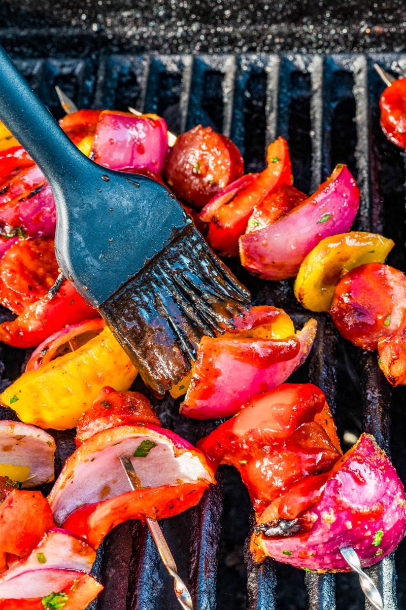 Vegetable skewers with red onions, tomatoes, and bell peppers are being brushed with sauce on a grill.