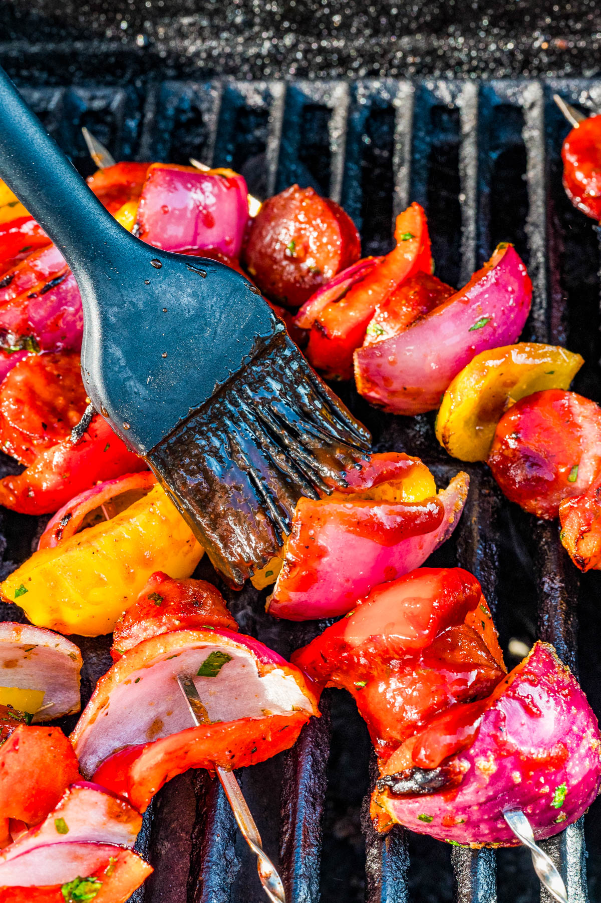 Vegetable skewers with red onions, tomatoes, and bell peppers are being brushed with sauce on a grill.