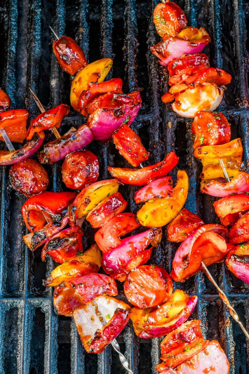 Colorful vegetable and tomato skewers grilling on a barbecue, with char marks visible on the vegetables and grill grates.