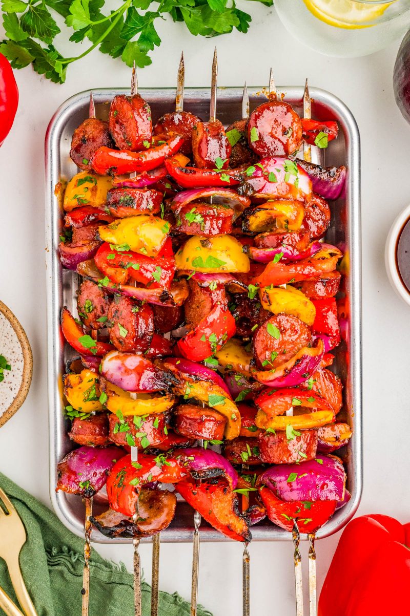 Metal skewers with grilled sausage, bell peppers, red onions, and herbs arranged on a metal tray, garnished with chopped parsley.