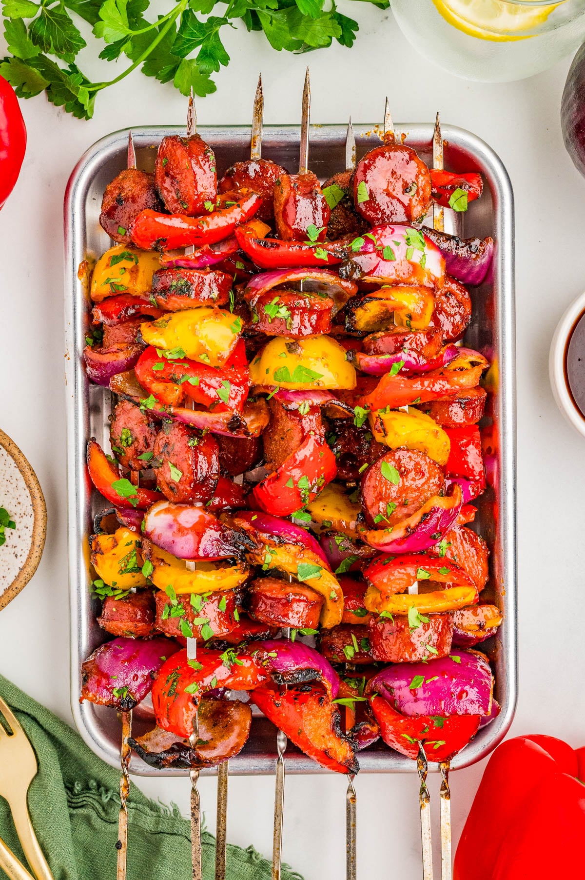 Metal skewers with grilled sausage, bell peppers, red onions, and herbs arranged on a metal tray, garnished with chopped parsley.