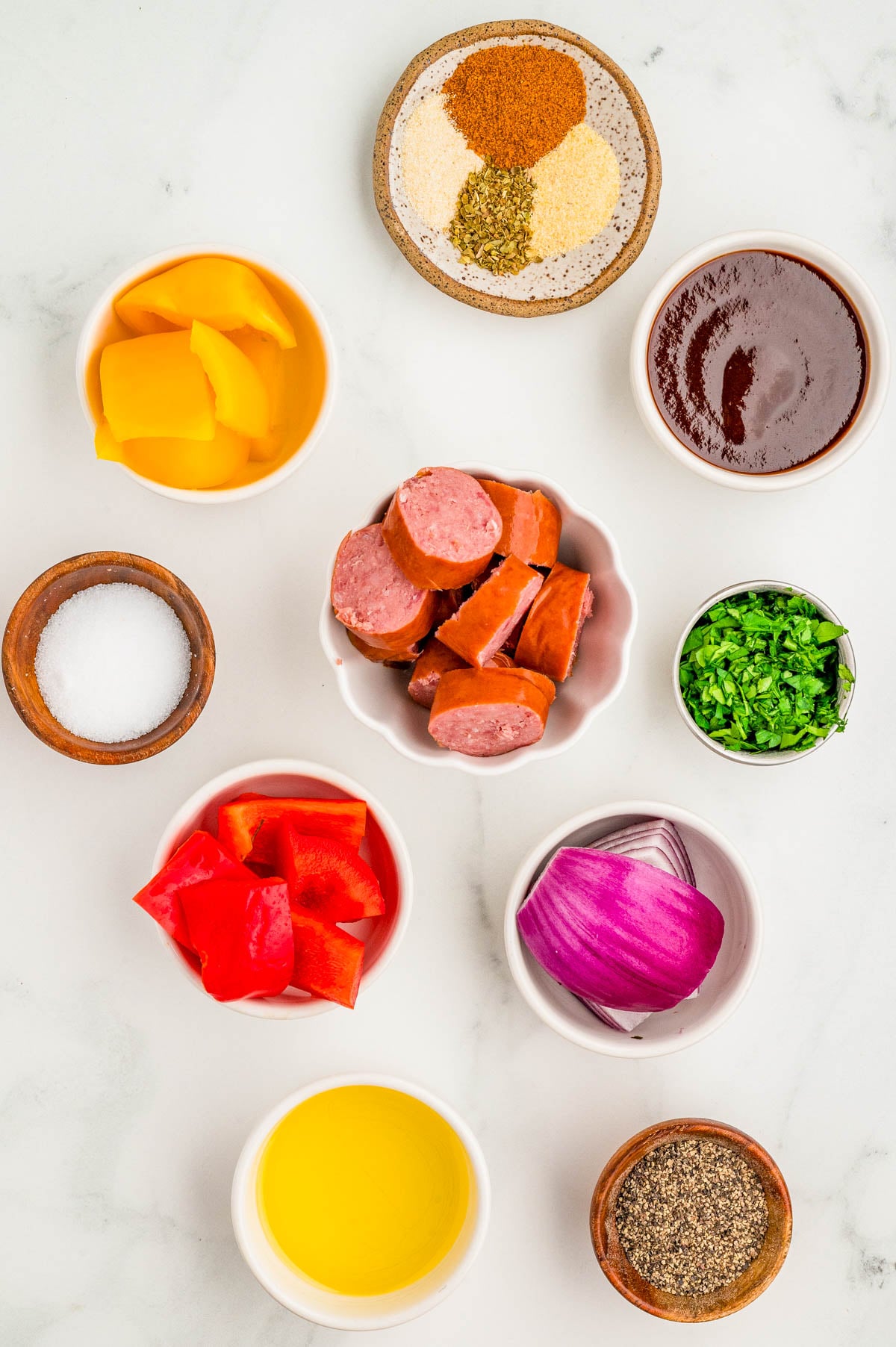 Assorted ingredients in bowls on a white surface, including sausage slices, chopped red and yellow bell peppers, red onion, seasonings, herbs, oil, barbecue sauce, salt, and pepper.