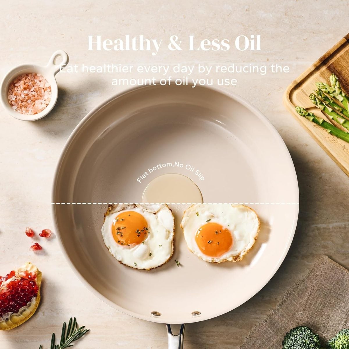 A beige non-stick frying pan with two sunny-side-up eggs sits on a countertop with pink salt, asparagus, broccoli, and a pomegranate nearby. Text promotes healthy, low-oil cooking.