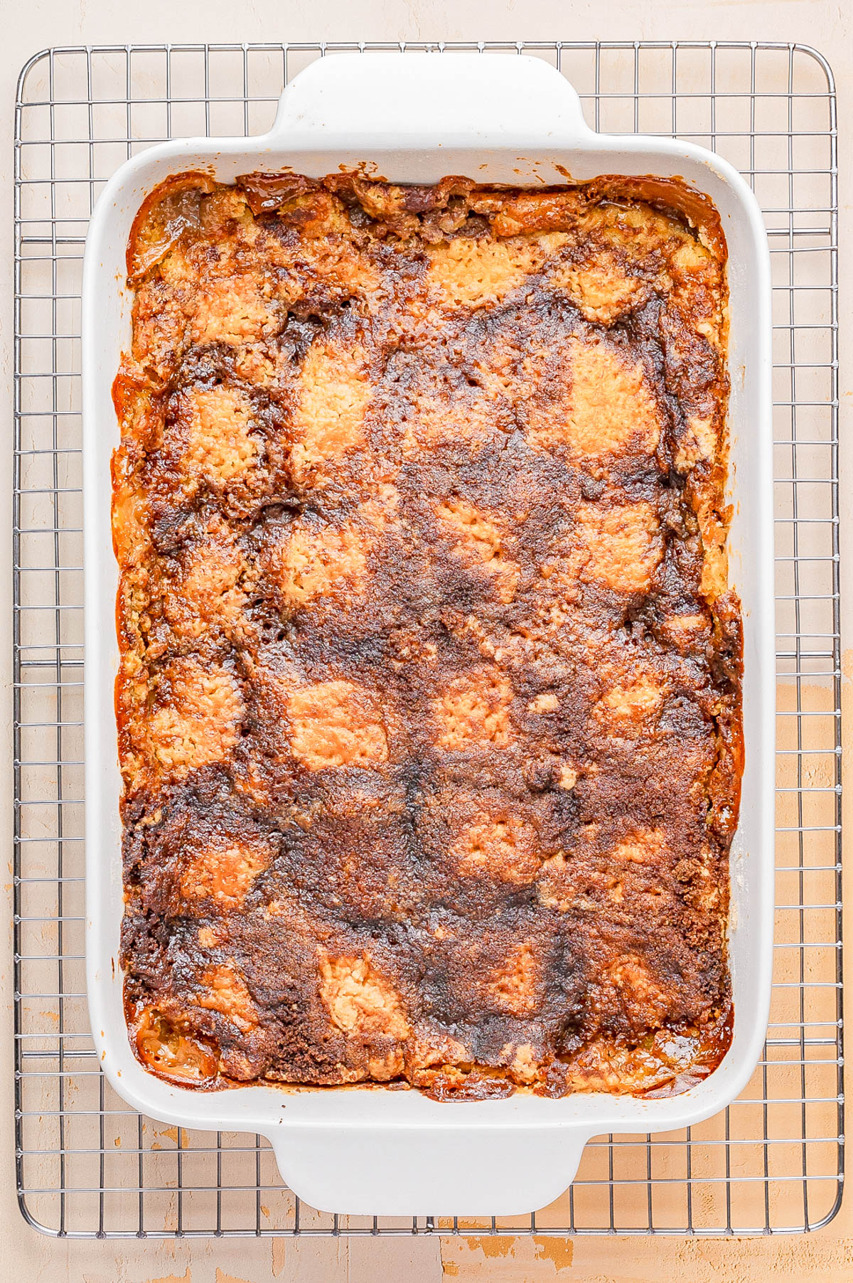 A baked cobbler with a golden-brown, slightly uneven crust in a white rectangular dish sits on a cooling rack.