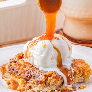 A serving of dessert topped with a scoop of vanilla ice cream, being drizzled with caramel sauce, is plated in front of a glass of milk.