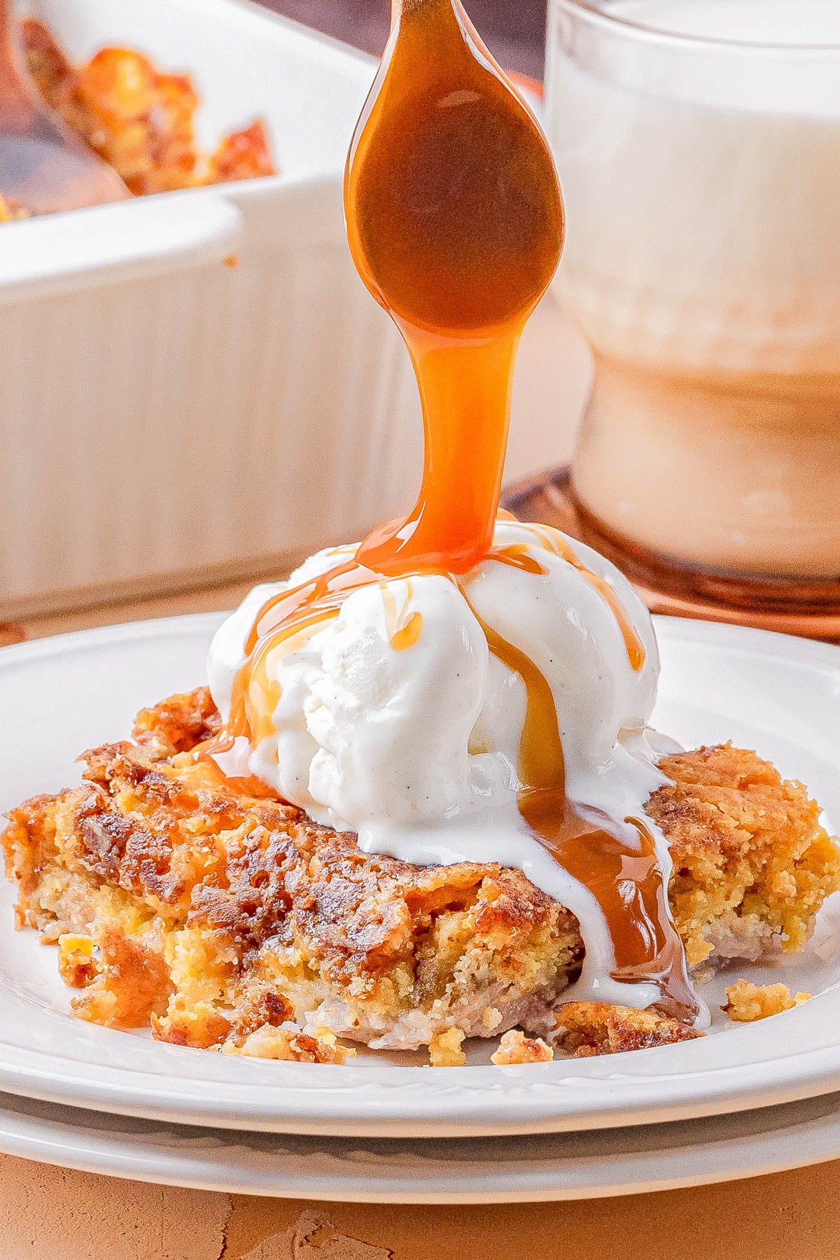 A serving of dessert topped with a scoop of vanilla ice cream, being drizzled with caramel sauce, is plated in front of a glass of milk.