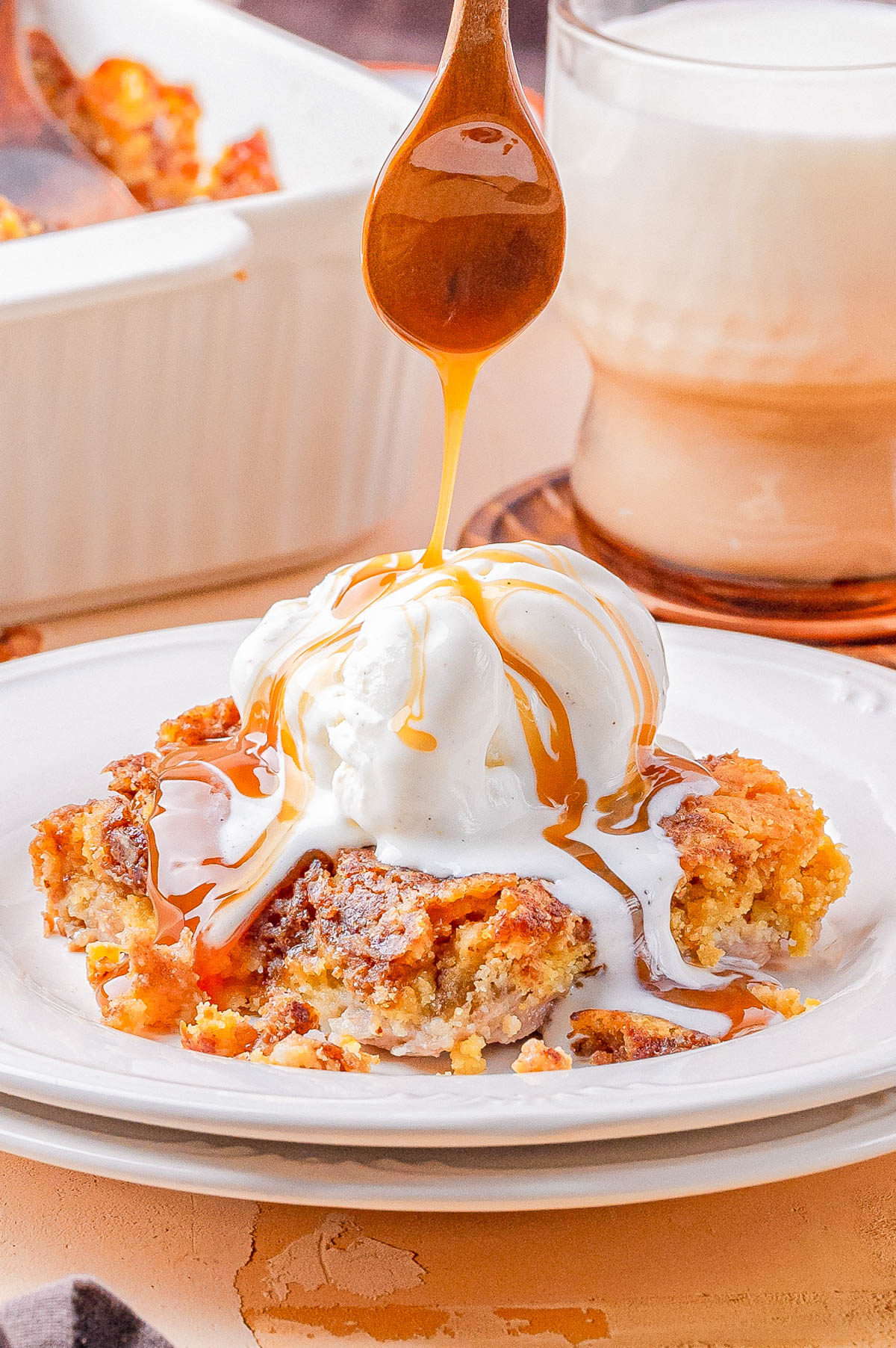 A dessert topped with a scoop of vanilla ice cream is drizzled with caramel sauce from a spoon. A glass of a creamy beverage and a baking dish are in the background.