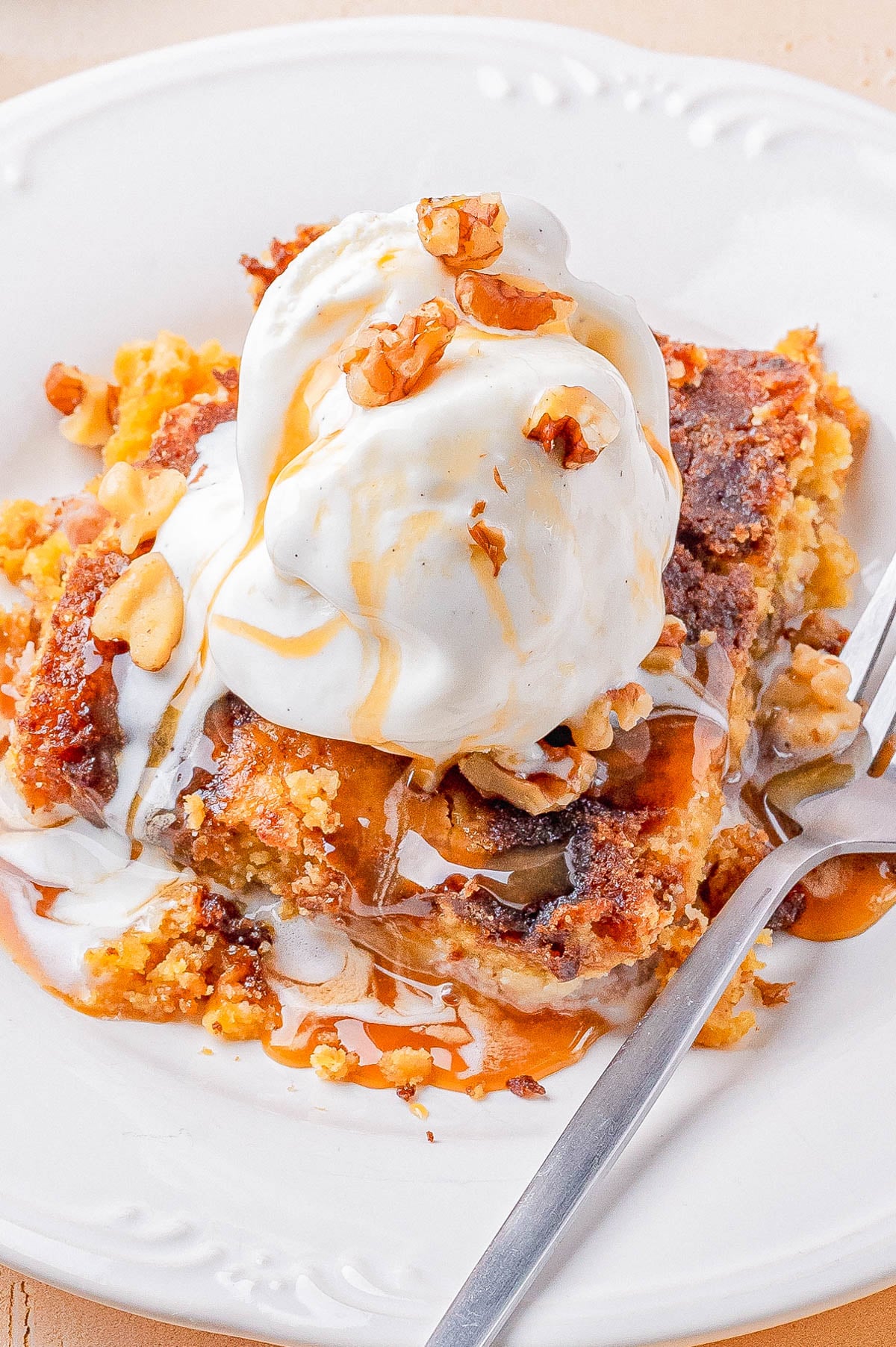 A slice of cake topped with a scoop of vanilla ice cream, chopped walnuts, and drizzled with syrup on a white plate with a fork.