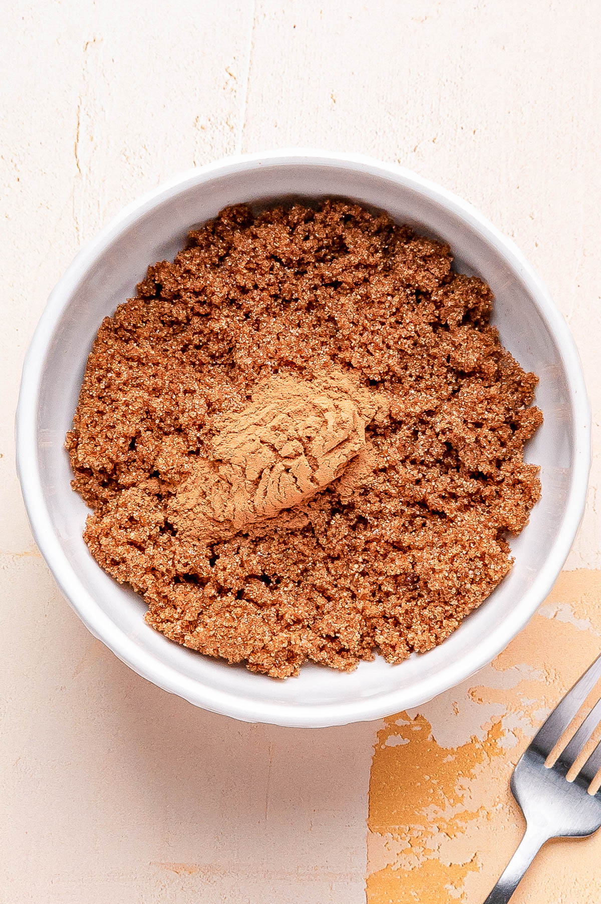 A white bowl filled with brown sugar and a scoop of ground cinnamon on top, sitting on a light-colored surface next to a metal fork.