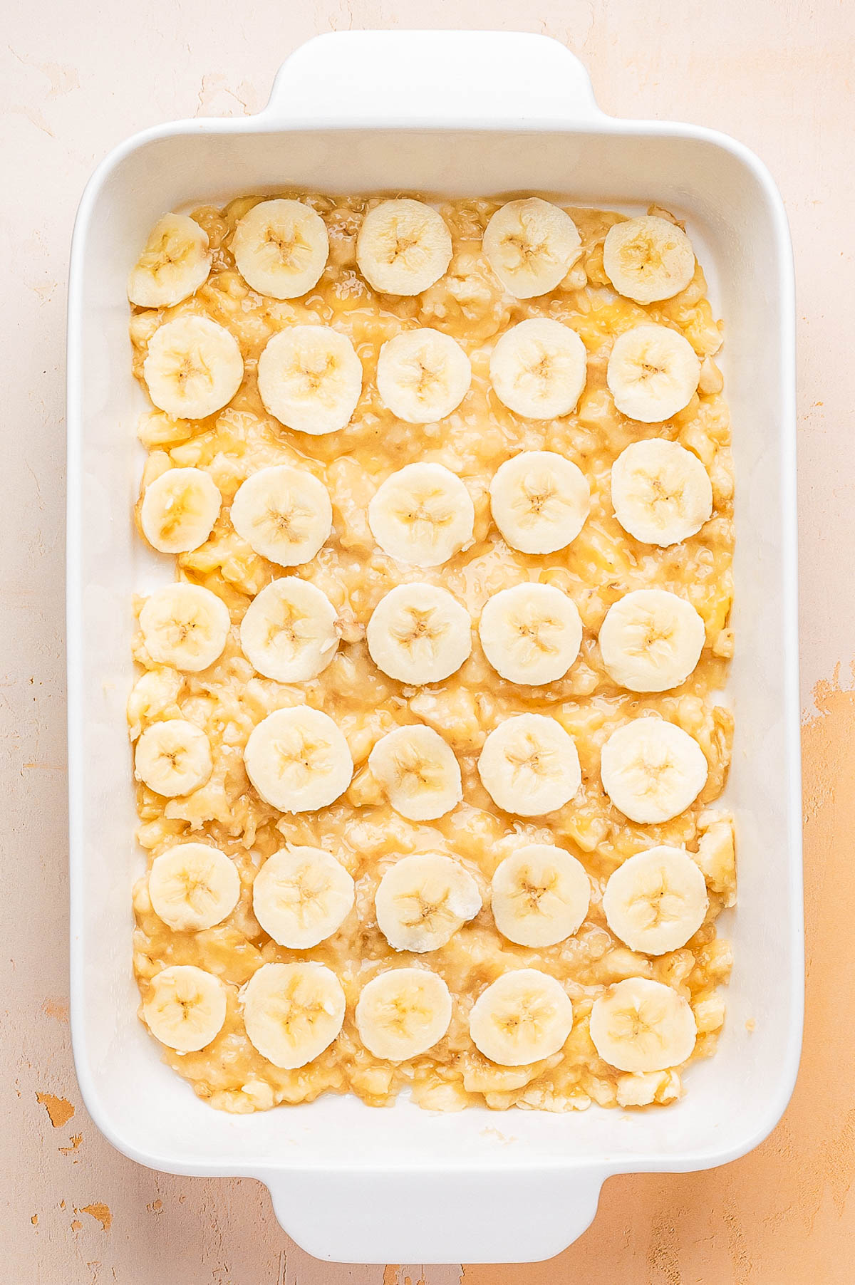 A white baking dish containing a layer of mashed bananas topped with evenly arranged banana slices.