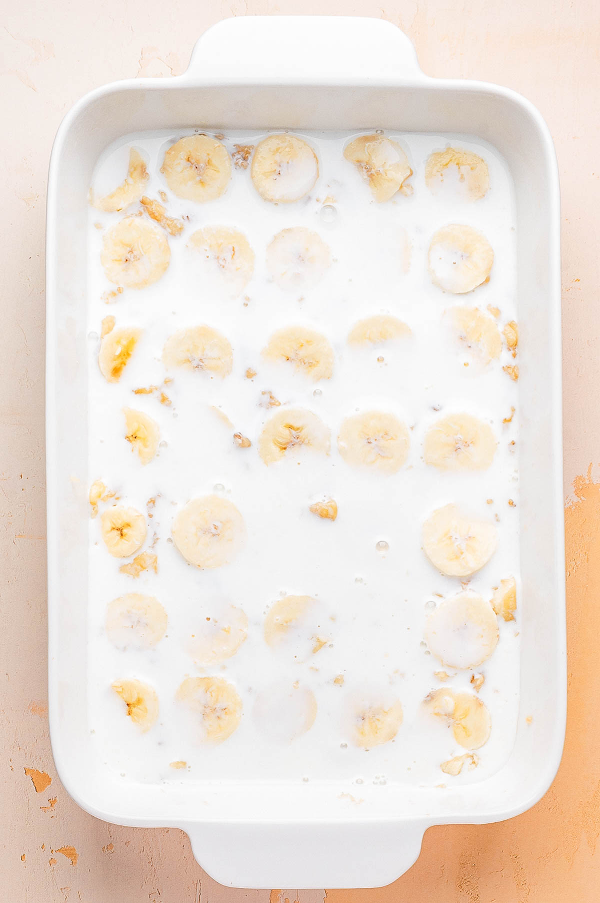A white baking dish filled with banana slices and covered in a milky liquid, viewed from above on a light-colored surface.