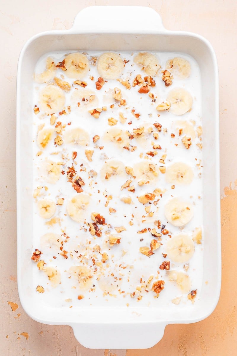 A white baking dish filled with sliced bananas, chopped walnuts, and milk.