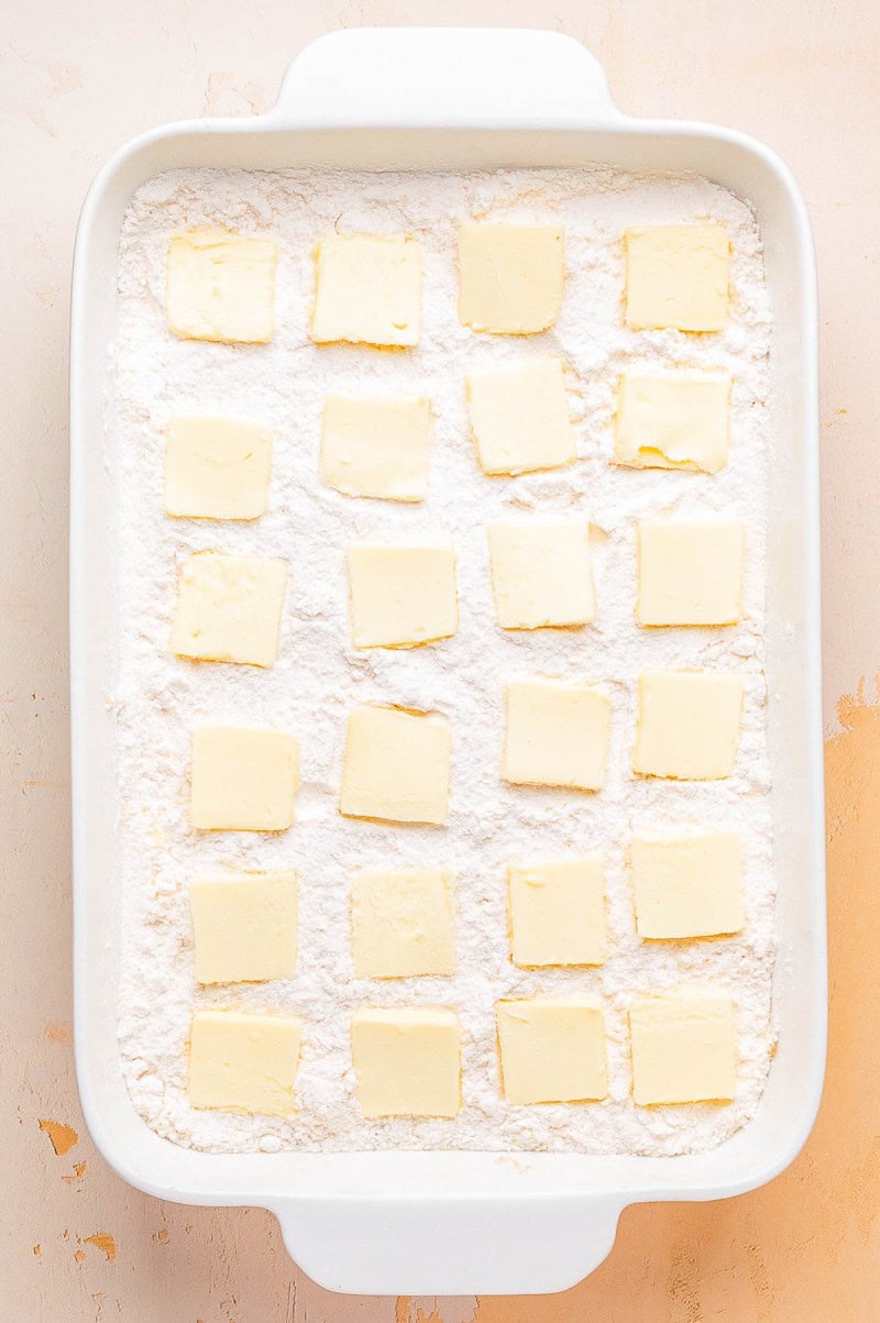 A white baking dish filled with a layer of white powder, topped with evenly spaced square slices of butter.